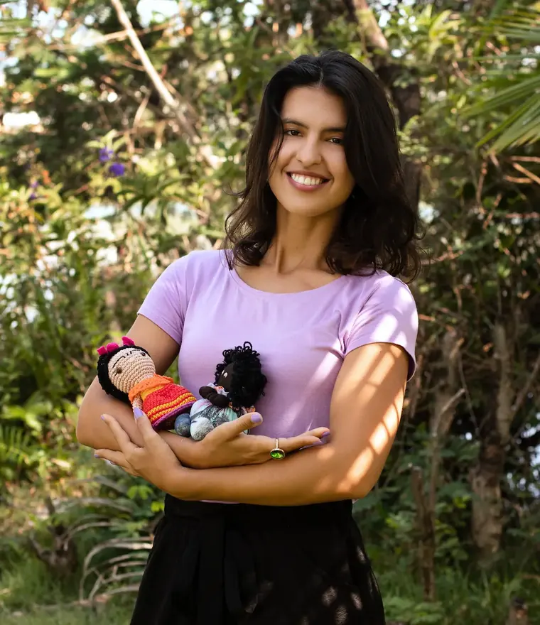 Lua Lordelo sorrindo ao lado de bonecas de crochê que fez, em um jardim